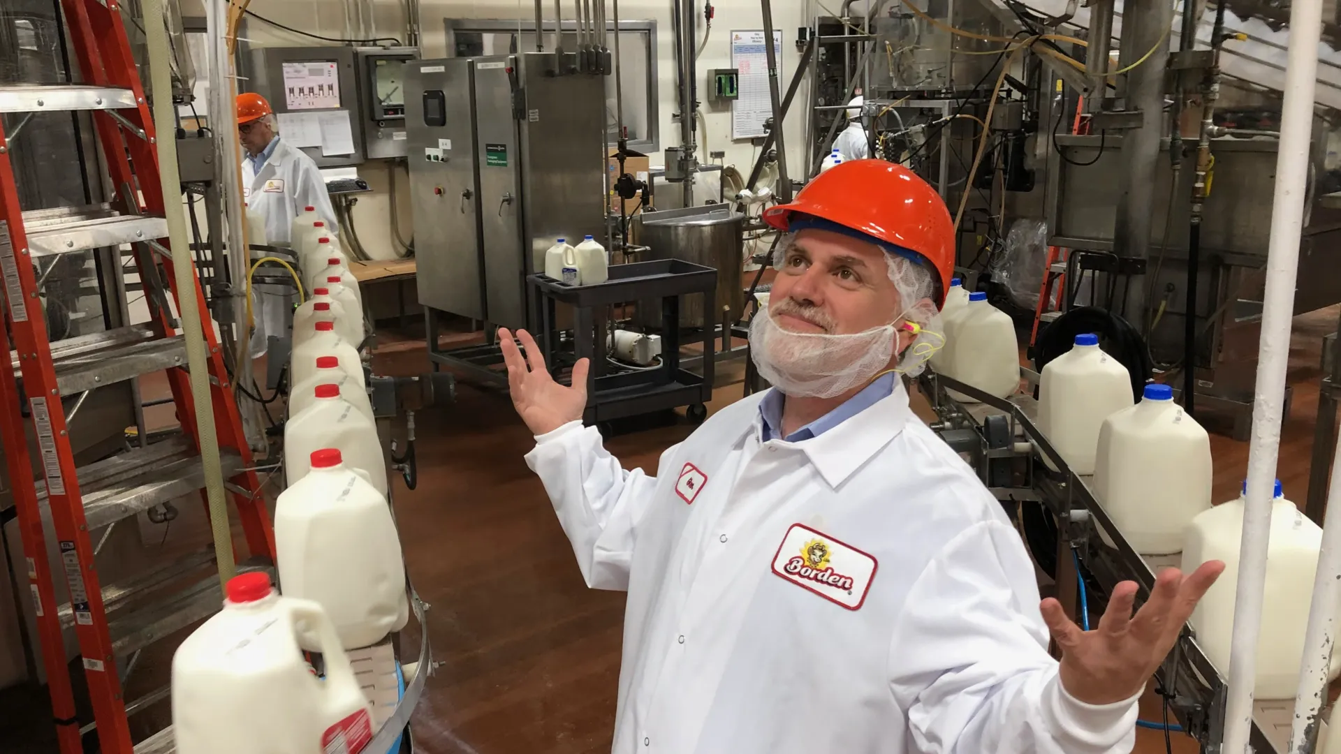 Tony Sarsam in a hairnet and Borden smock gesturing on a milk bottling line as filled gallon jugs move along the conveyor